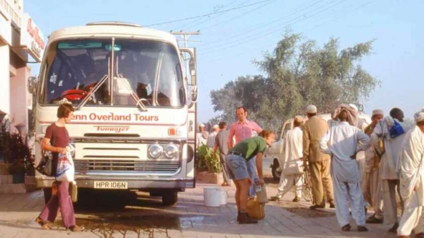 A white tour bus labeled "Penn Overland Tours" is parked on a street. A group of people, some in traditional South Asian clothing, stand nearby while a man fills a container with water and a woman walks by holding bags.