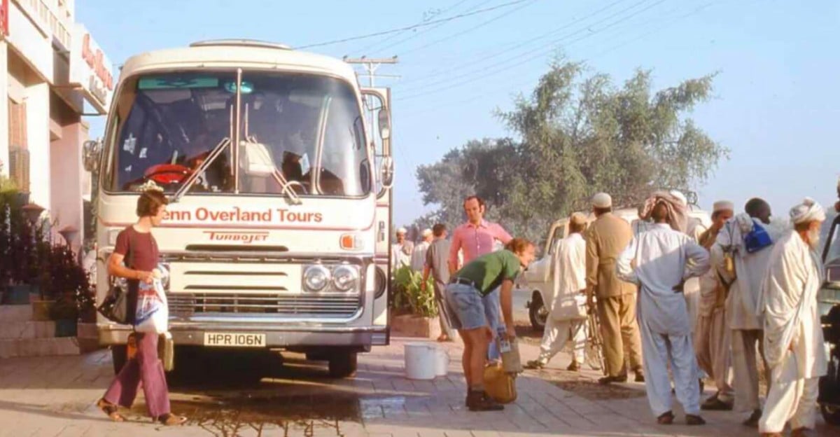 A white tour bus labeled "Penn Overland Tours" is parked on a street. A group of people, some in traditional South Asian clothing, stand nearby while a man fills a container with water and a woman walks by holding bags.