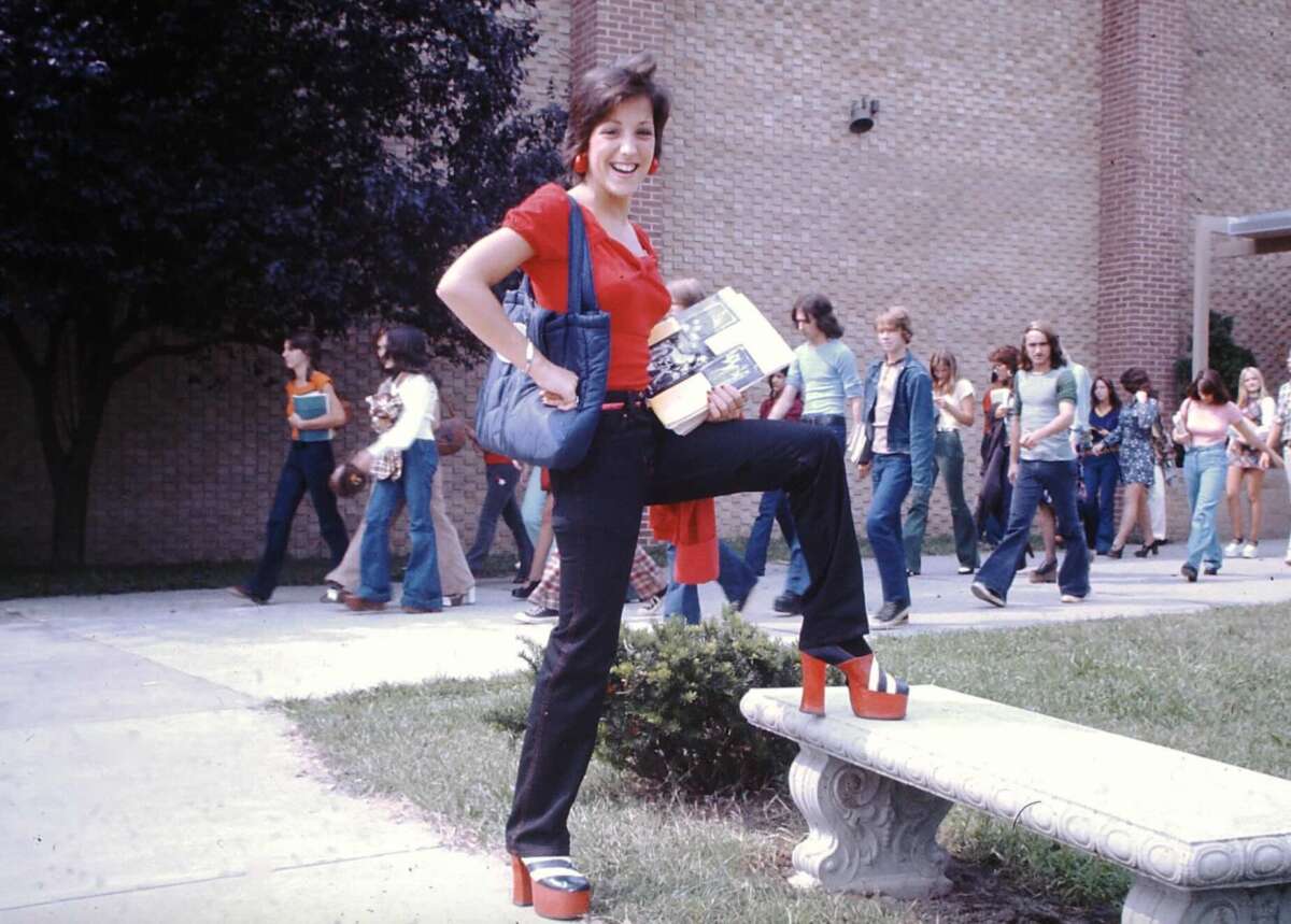 A smiling young woman in a red top and platform shoes poses with one foot on a stone bench, holding books and a bag, as students walk behind her outside a brick school building.