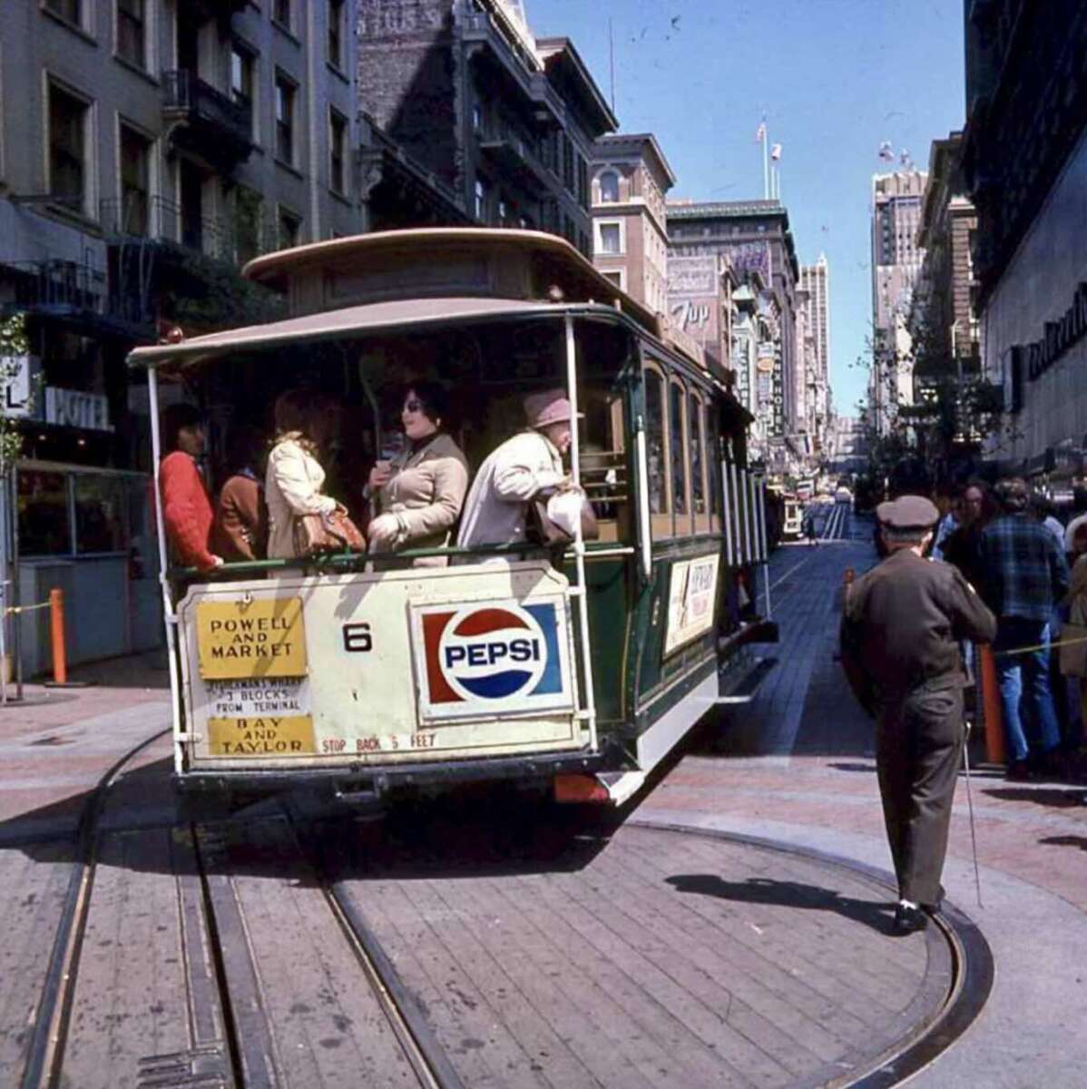 A vintage San Francisco cable car filled with passengers travels down a city street. Shops and buildings line the road, and a man stands near the tracks in the foreground. A large Pepsi ad is displayed on the cable car.