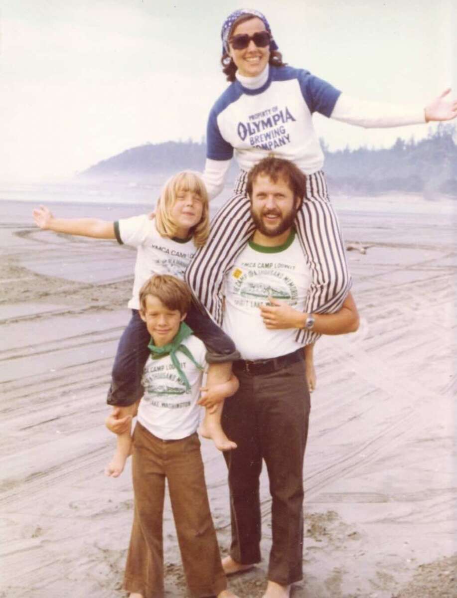 Four people pose on a sandy beach. A man stands with a child on his shoulders and another child beside him. Another adult stands behind, arms outstretched. All are smiling and dressed in casual clothing. Trees and ocean are in the background.