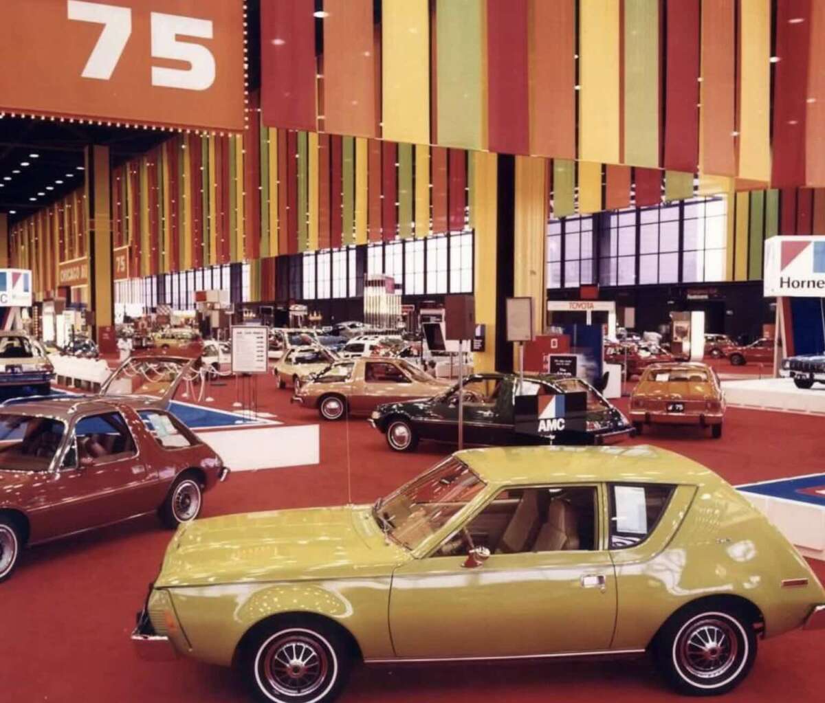 A vintage car show with brightly colored cars and bold red, yellow, and orange hanging decor. The number "75" is displayed above, indicating the year or event, and several classic cars are arranged on a red carpet.