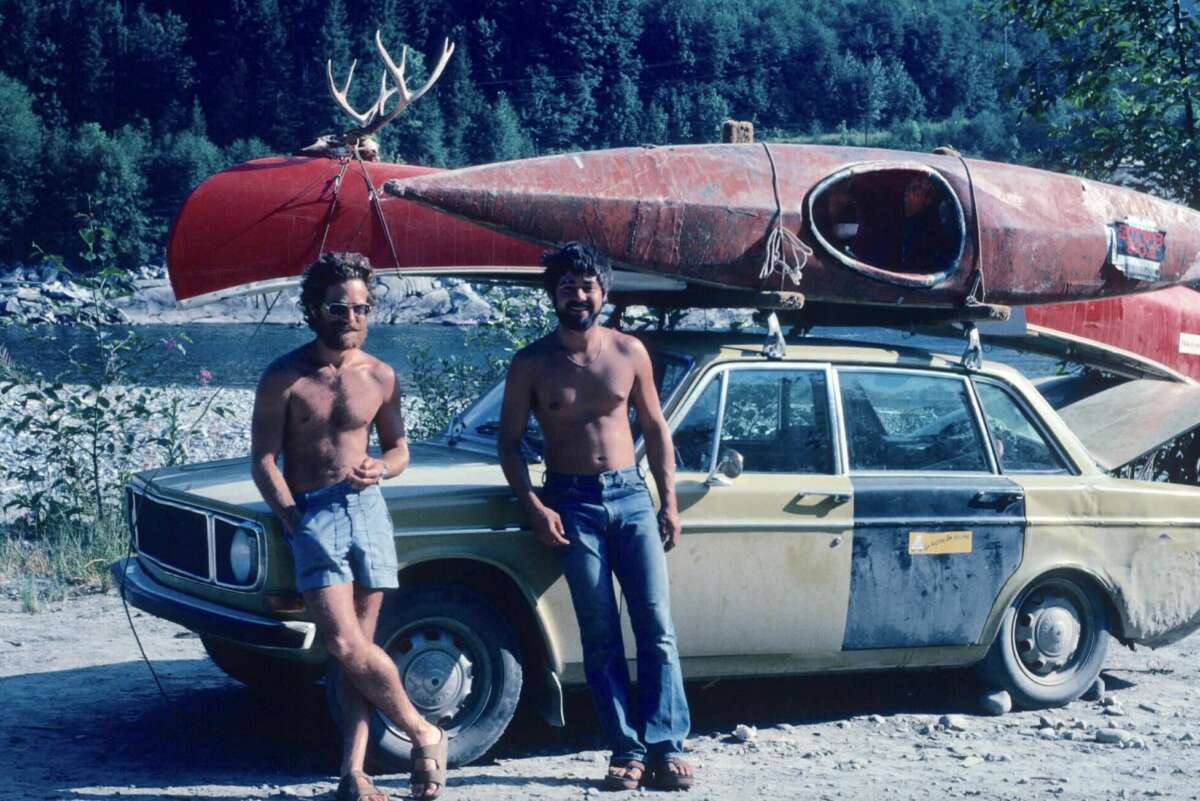 Two men with beards, shirtless, lean against an old car with a red kayak and antlers strapped to the roof; a river and forested hillside are in the background on a sunny day.