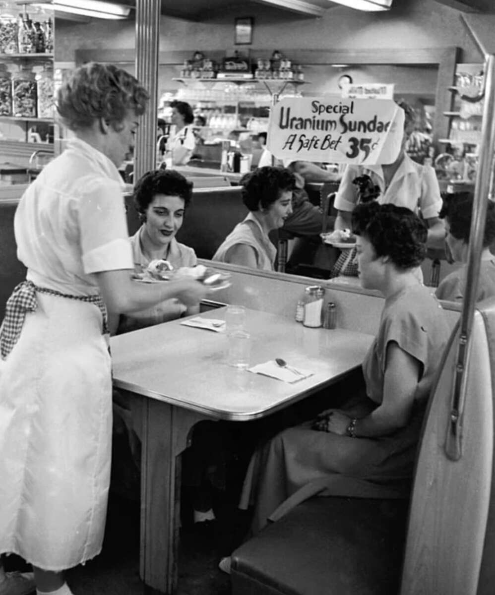Black-and-white photo of women sitting at a diner booth being served by a waitress. A sign behind them advertises a “Special Uranium Sundae - A Safe Bet 35¢.” Shelves of products are visible in the background.