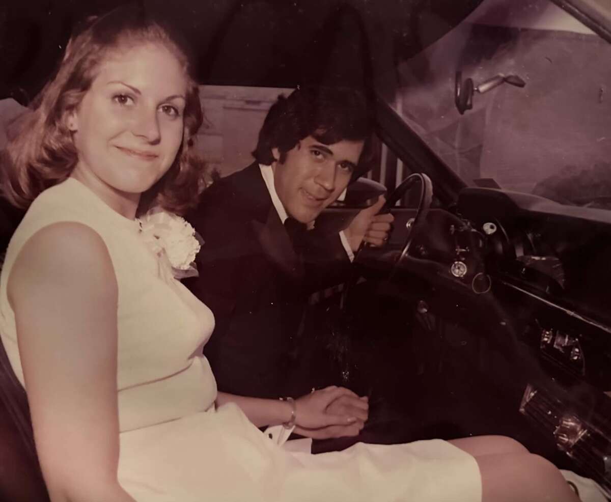 A young woman in a white dress and a man in a suit sit smiling in the front seats of a car. The man is in the driver's seat, holding the steering wheel, while the woman looks at the camera.