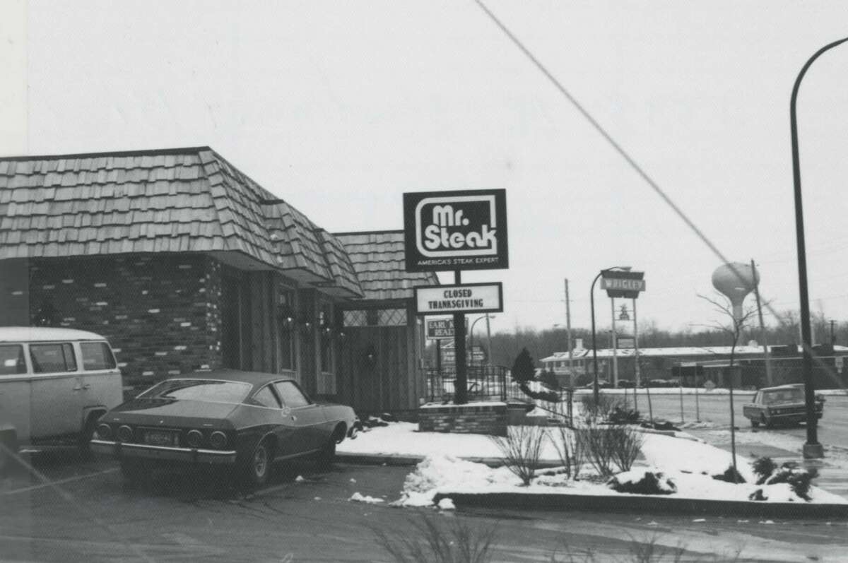 A black-and-white photo of a Mr. Steak restaurant with snow on the ground, a parked vintage car, and a van. The restaurant sign reads "Mr. Steak" and "Closed Thanksgiving." Nearby are street signs and a water tower.