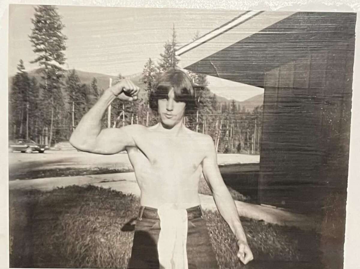 A young man with medium-length hair poses shirtless outdoors, flexing his right arm in a muscle pose. Trees, mountains, and a building are visible in the background. The photo is in black and white.