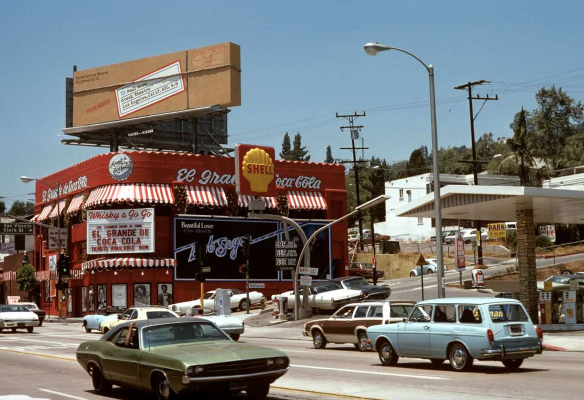 A busy street scene from the 1970s with vintage cars driving past a red nightclub called Whisky a Go Go and a Shell gas station. A large billboard and various signs are visible against a sunny backdrop.