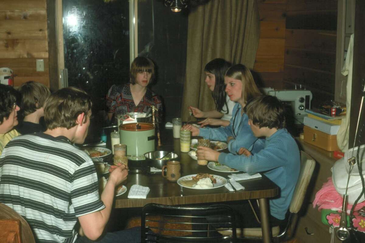 A group of young people sit around a dining table in a cozy, wood-paneled room, eating a meal together. The table is set with food, drinks, and condiments, and a window and curtain are visible in the background.