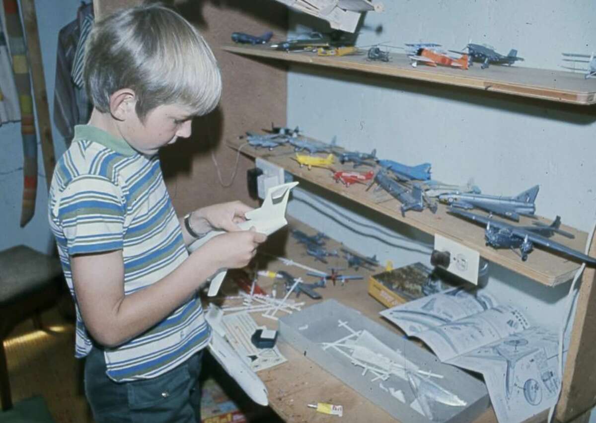A young boy in a striped shirt assembles model airplanes at a desk filled with various completed aircraft models, plastic parts, tools, and instruction sheets.