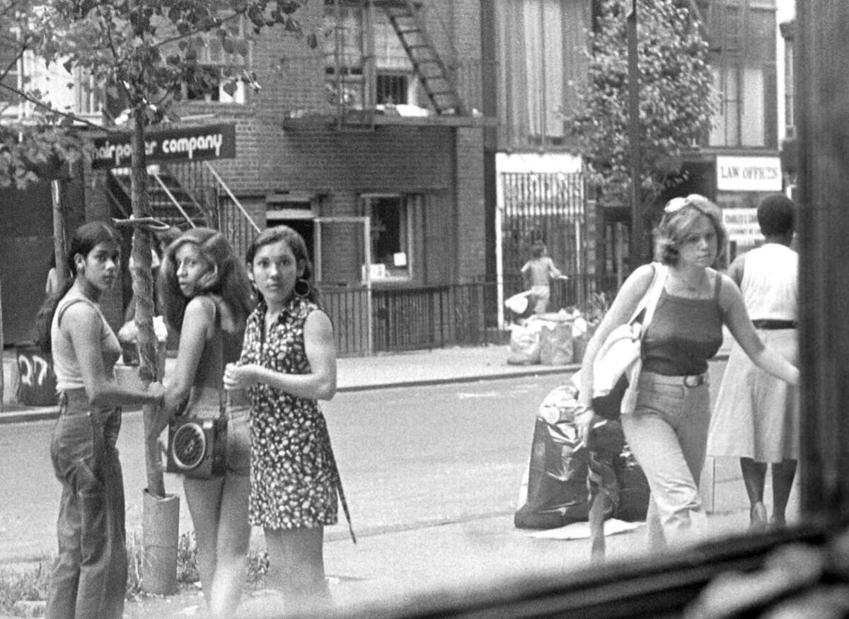 Black and white photo of four young women standing on a city sidewalk, two looking toward the camera. In the background, people walk past storefronts, trash bags, and a fire escape. The scene captures an everyday urban moment.