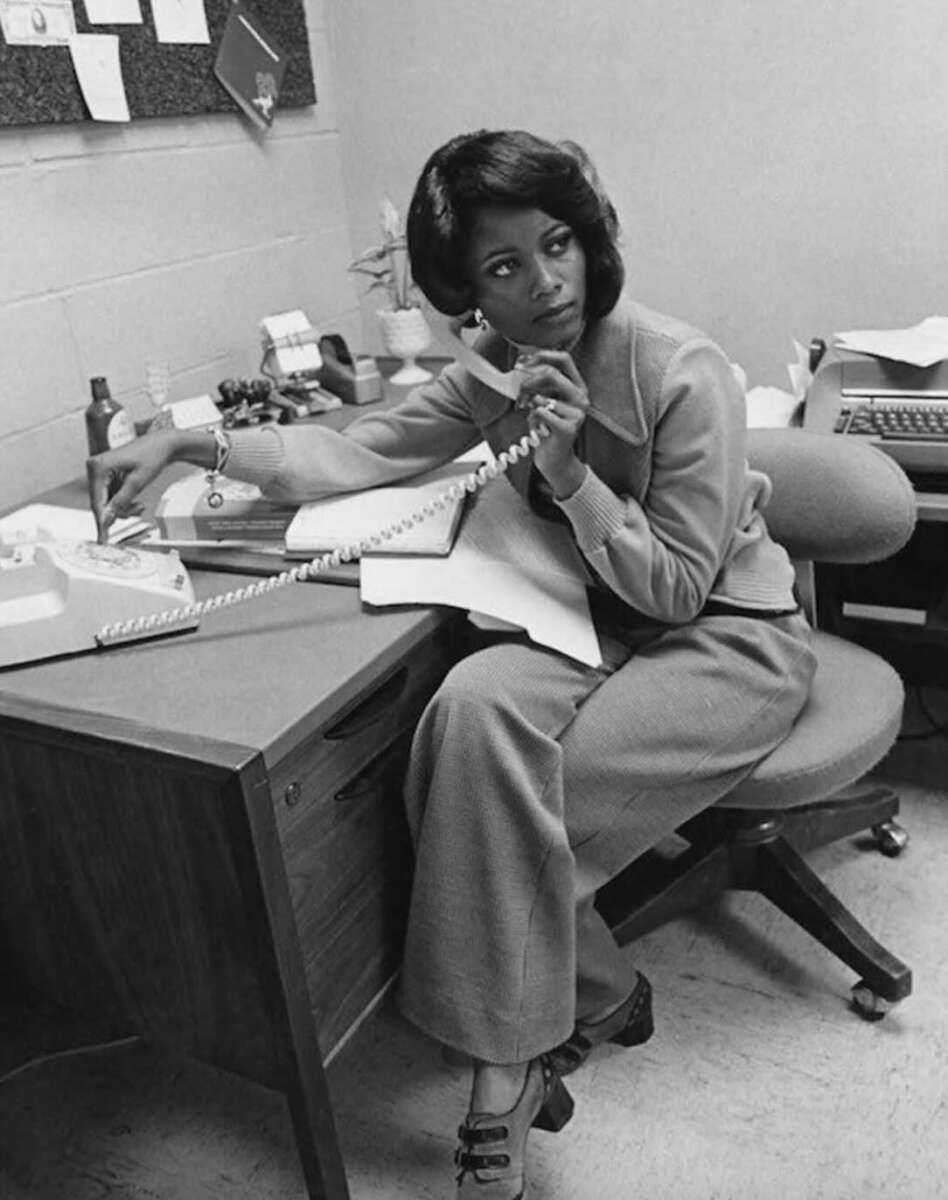 A woman in 1970s-style clothing sits at an office desk, talking on a corded phone while writing on paper. She appears focused, with office supplies, documents, and a typewriter visible on the desk.