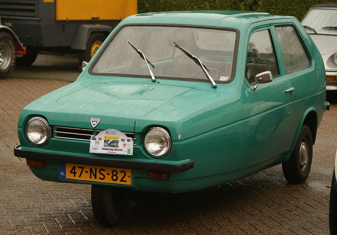 A green three-wheeled Reliant Robin car is parked on a wet brick road, with raindrops on its surface and a Dutch license plate reading "47-NS-82". A sign is attached to the front grille.