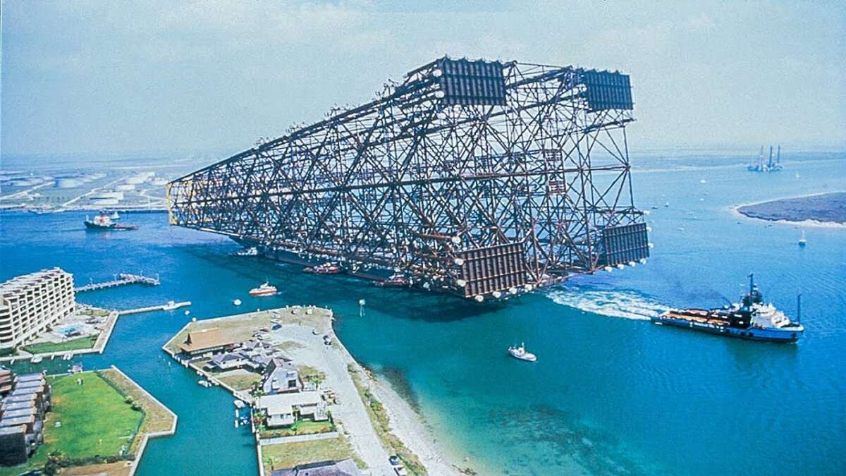 A massive steel oil platform jacket structure is being transported by tugboats through blue waters near a coastal area with buildings, greenery, and a distant shoreline under a clear sky.