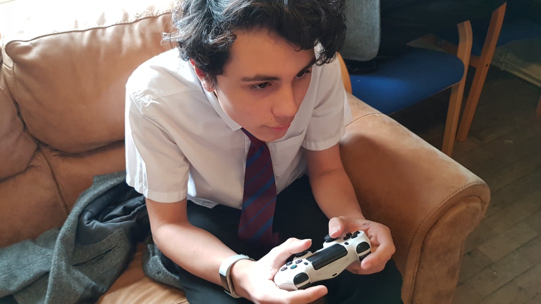 A young person in a school uniform and striped tie sits on a tan couch, holding a video game controller and focusing intently ahead. A jacket is draped beside them on the couch.