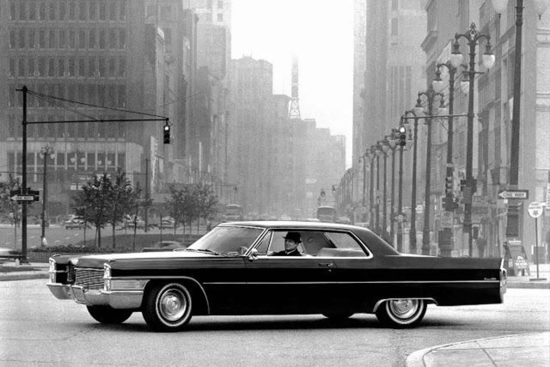 A classic black car with a driver wearing a hat is stopped at an intersection in a city with tall buildings and streetlights, giving a vintage urban atmosphere.