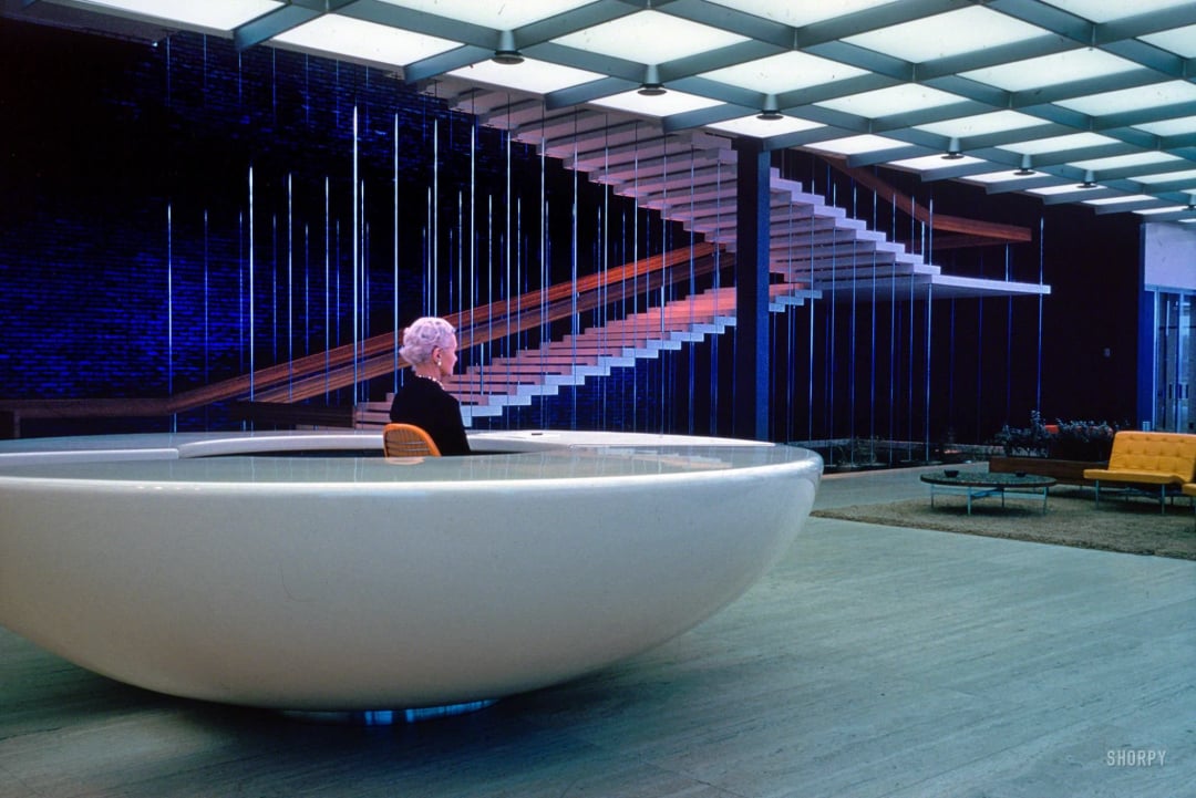 A woman with short white hair sits at a futuristic, white oval reception desk in a spacious, modern lobby with a floating staircase and a grid of ceiling lights.