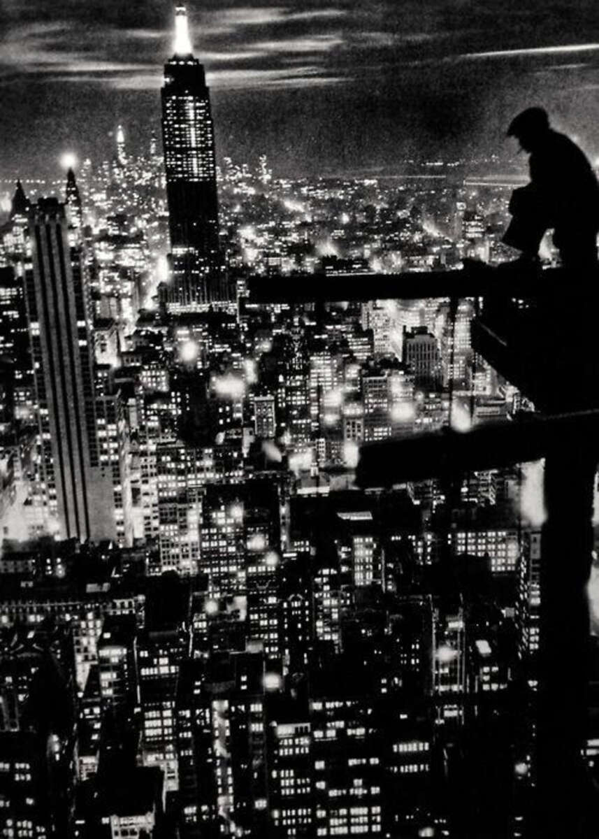 A black-and-white photo of New York City at night, with a construction worker silhouetted on steel beams high above the city and the Empire State Building illuminated in the background.