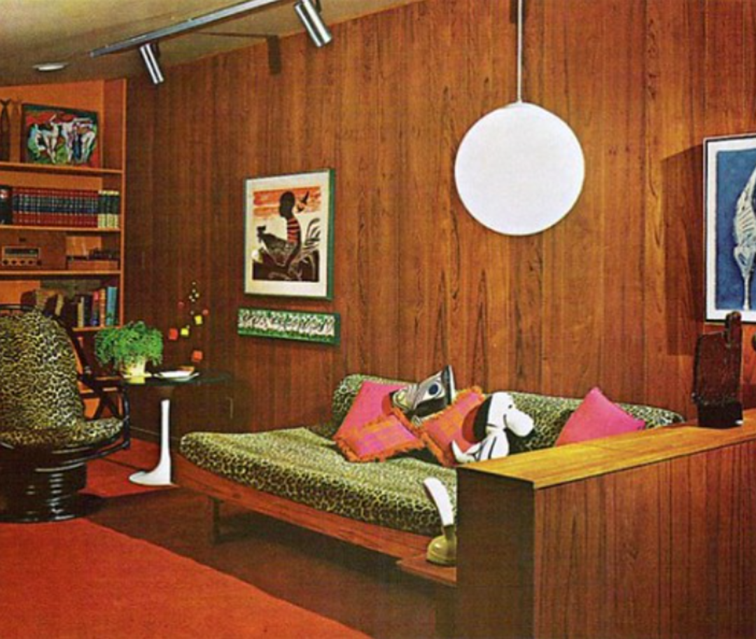 A retro living room with wood-paneled walls, an animal-print sofa, vibrant pillows, a globe pendant light, bookshelf, art prints, and a mid-century modern chair with a matching animal-print pattern.