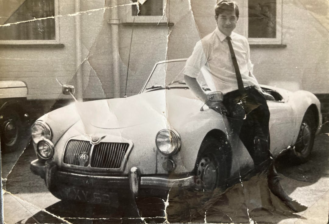 A young man in a white shirt and tie sits on the hood of a vintage convertible car, smiling. The black-and-white photo appears old and is visibly creased and worn. A building is seen in the background.