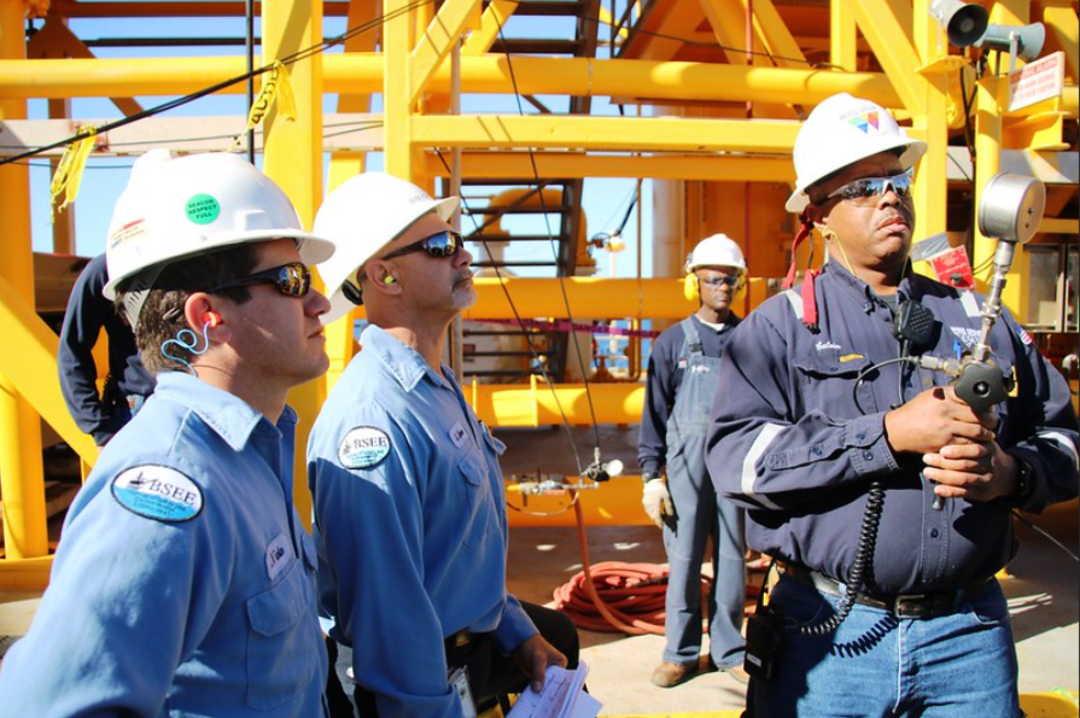 Four workers wearing hard hats, safety glasses, and blue uniforms stand on an industrial site with yellow metal structures, appearing focused and engaged in a discussion or safety inspection.