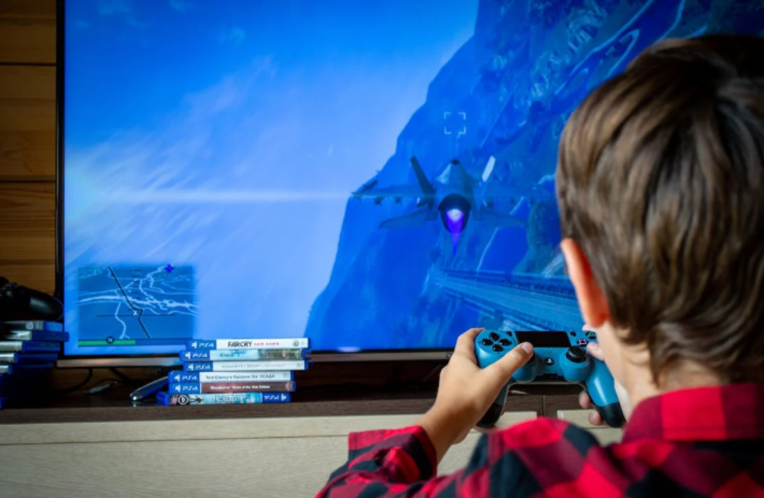 A child holding a PlayStation controller plays a flight video game on a large TV screen. Several video game cases are stacked on the table beside the child. The room has wooden walls and a cozy atmosphere.