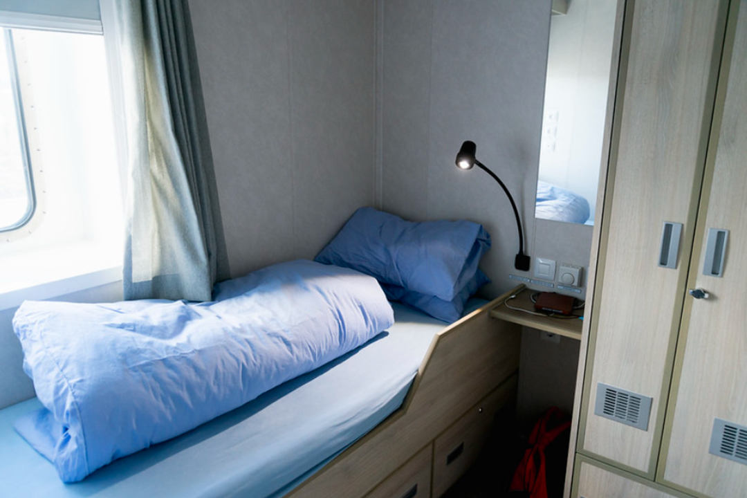 A small, tidy room with a single bed covered in blue bedding, a bedside table with a lamp, and a wardrobe. Daylight comes through a window partially covered by curtains.