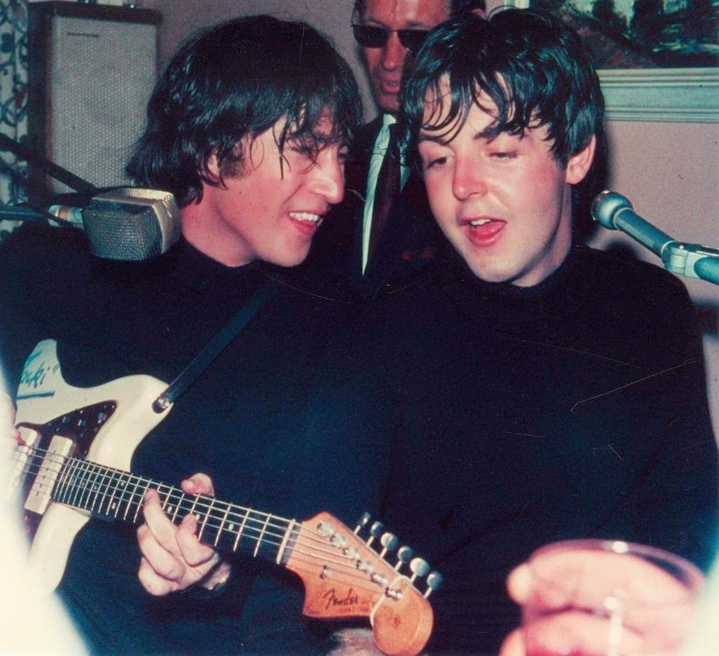 Two young men with shaggy hair, wearing black turtlenecks, sit closely together singing into microphones. One plays an electric guitar while smiling at the other. Another man stands behind them, partially visible.