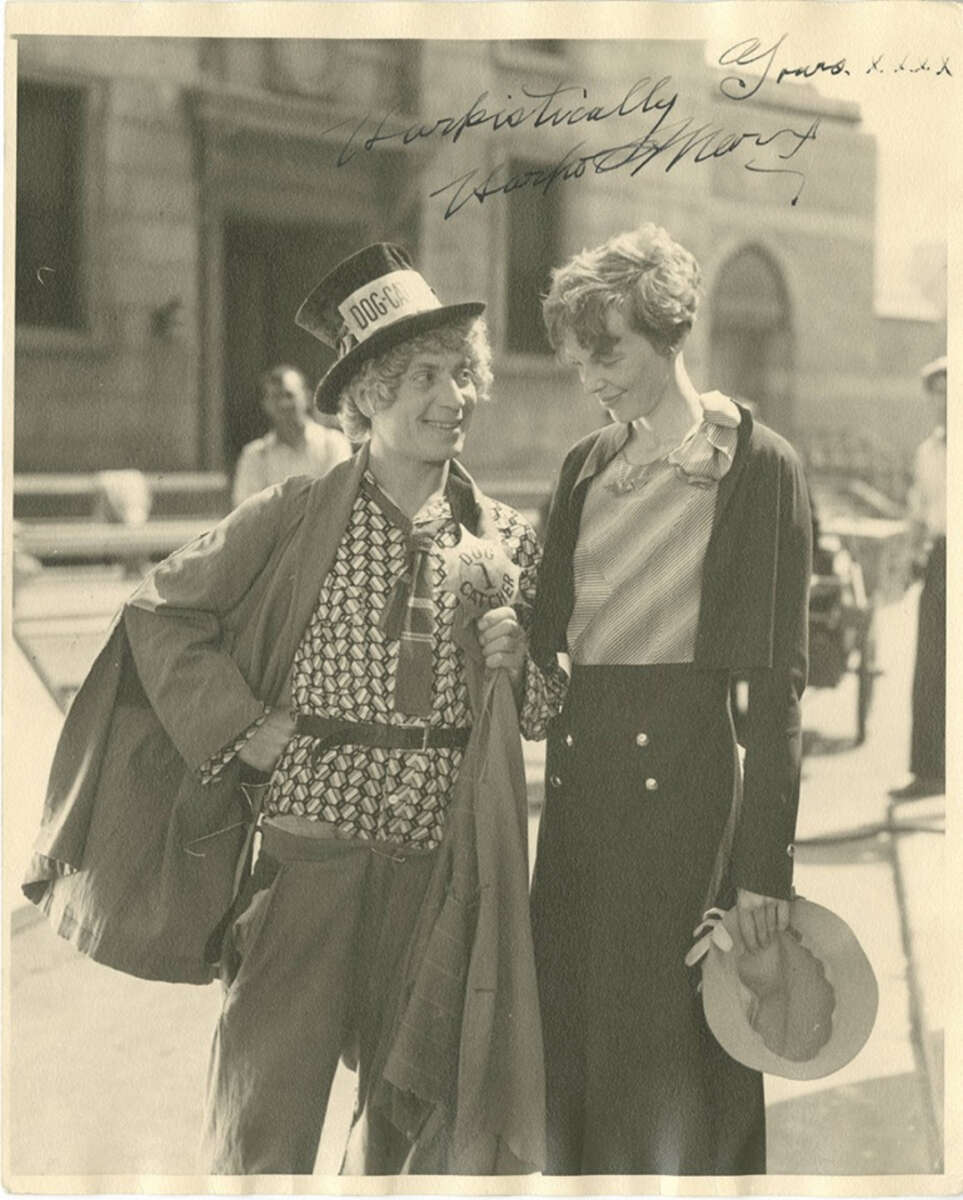 A person in whimsical clothing and a "DOGCATCHER" hat smiles at a woman in a dress holding a hat, both standing outdoors and looking at each other warmly. The photo has handwritten signatures at the top.