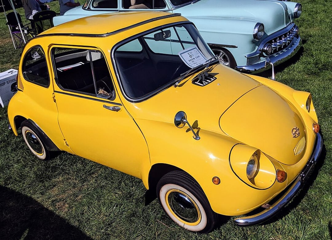 A bright yellow vintage Subaru 360 car with round headlights is parked on grass at a car show, next to a light blue classic car in the background. A sign is displayed on the dashboard inside the yellow car.