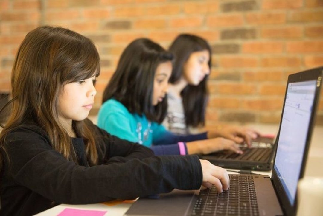 Three young girls sit side by side at a table, each focused on typing on their own laptops. The background features a brick wall, and the scene suggests a collaborative or educational setting.