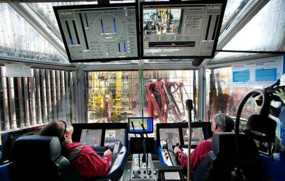 Two people in a control room operate equipment using multiple screens and control panels, overlooking a large industrial or drilling site visible through windows in front of them.