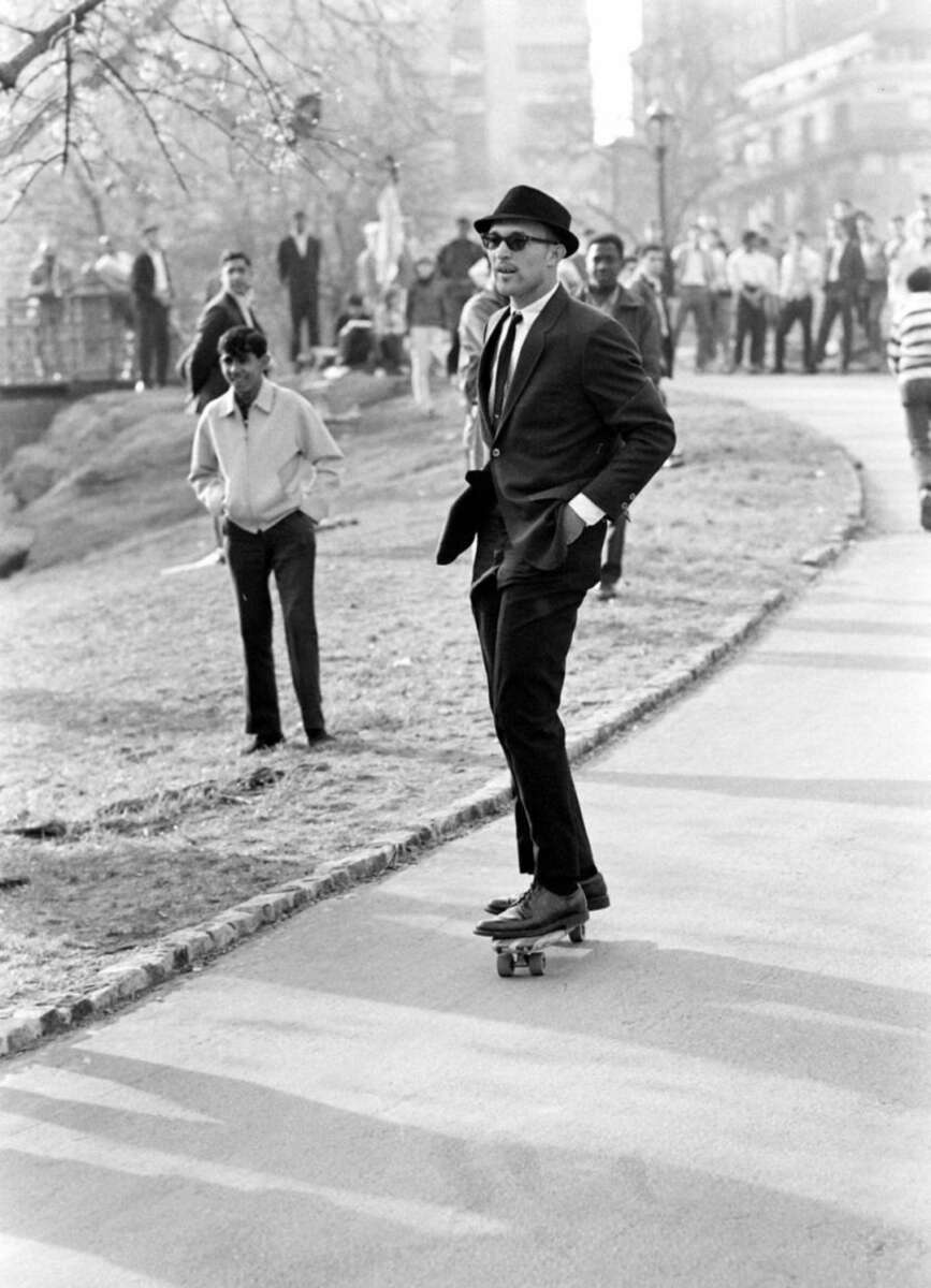 A man in a suit, tie, hat, and sunglasses rides a skateboard on a paved path in a park, with people watching in the background on a sunny day.