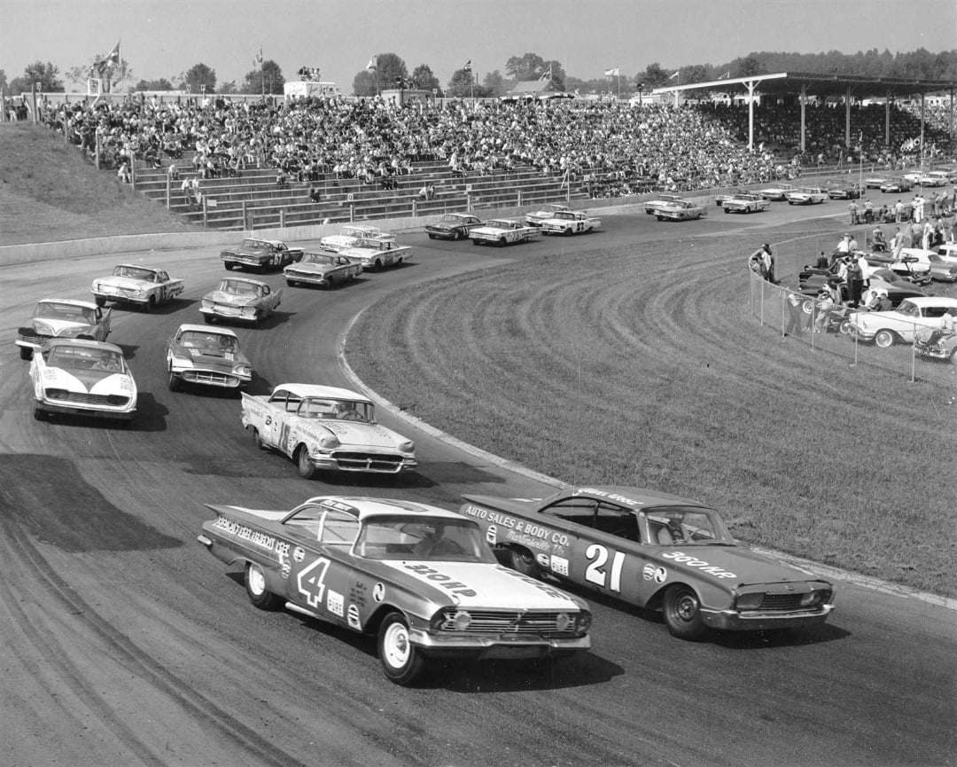 Black and white photo of a crowded car race on a curved track, with classic stock cars and grandstands full of spectators in the background. Two lead cars, numbered 4 and 21, are side by side at the front.