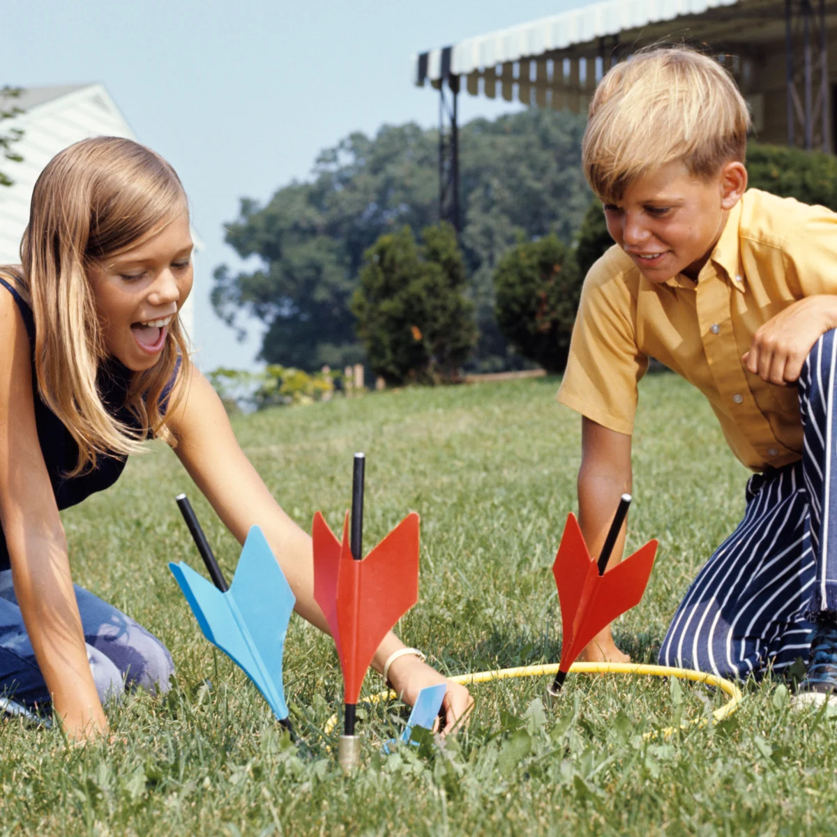 Two children, a girl and a boy, play lawn darts on green grass. Three large darts, two red and one blue, are stuck in the ground near a yellow ring. Both kids appear happy and focused on the game.