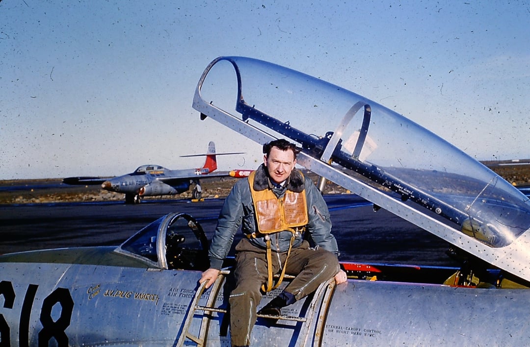 A man in a flight suit sits on the wing of a military jet with an open cockpit. Another jet is visible in the background on the tarmac under a clear blue sky.