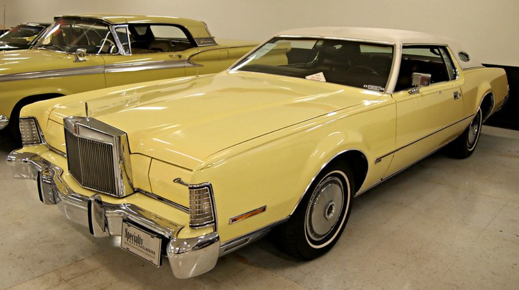 A vintage yellow Lincoln Continental Mark IV with a white roof on display indoors, featuring chrome detailing and classic styling, parked next to another classic car.