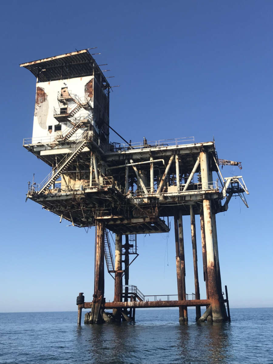 A rusty, abandoned offshore platform with a decaying structure stands above calm blue water under a clear sky. Stairs and railings are visible, and the platform appears weathered and unused.