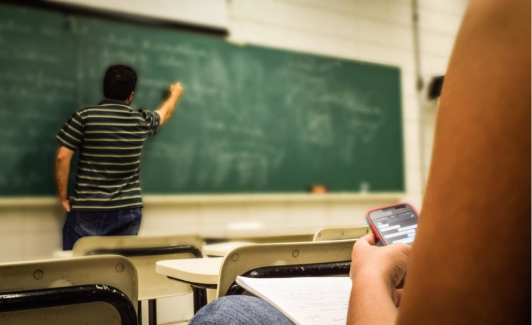 A person sits in a classroom using a smartphone while an instructor writes on a chalkboard at the front of the room. Desks and empty chairs are visible, creating a typical classroom setting.