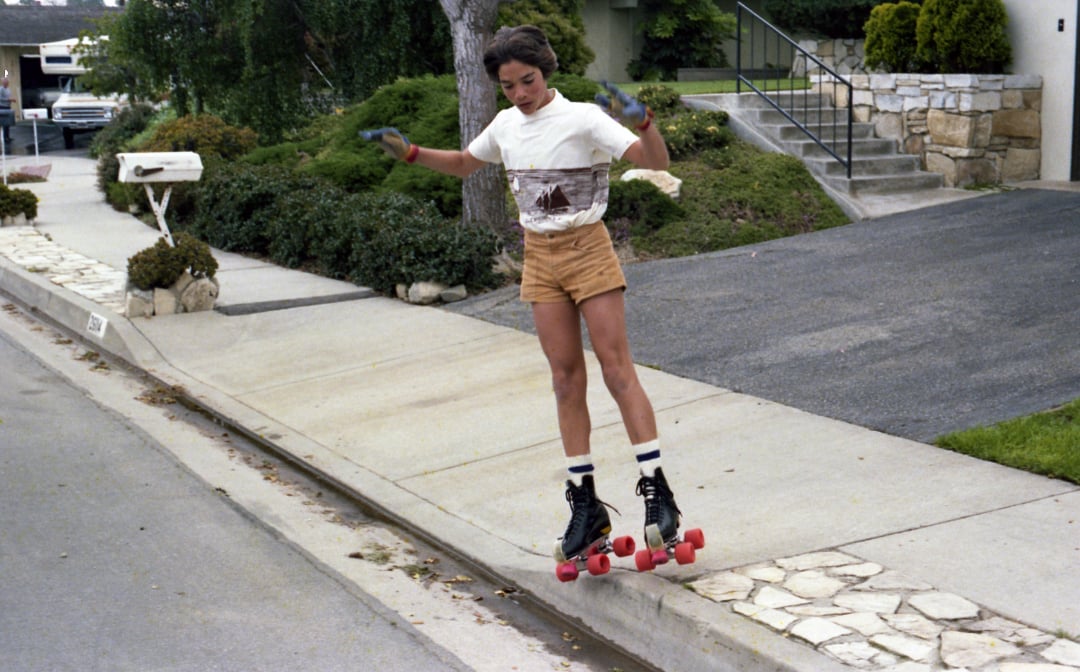 A person wearing vintage roller skates, shorts, and a white T-shirt is skating off a curb on a suburban street, with houses, greenery, and a white mailbox in the background.