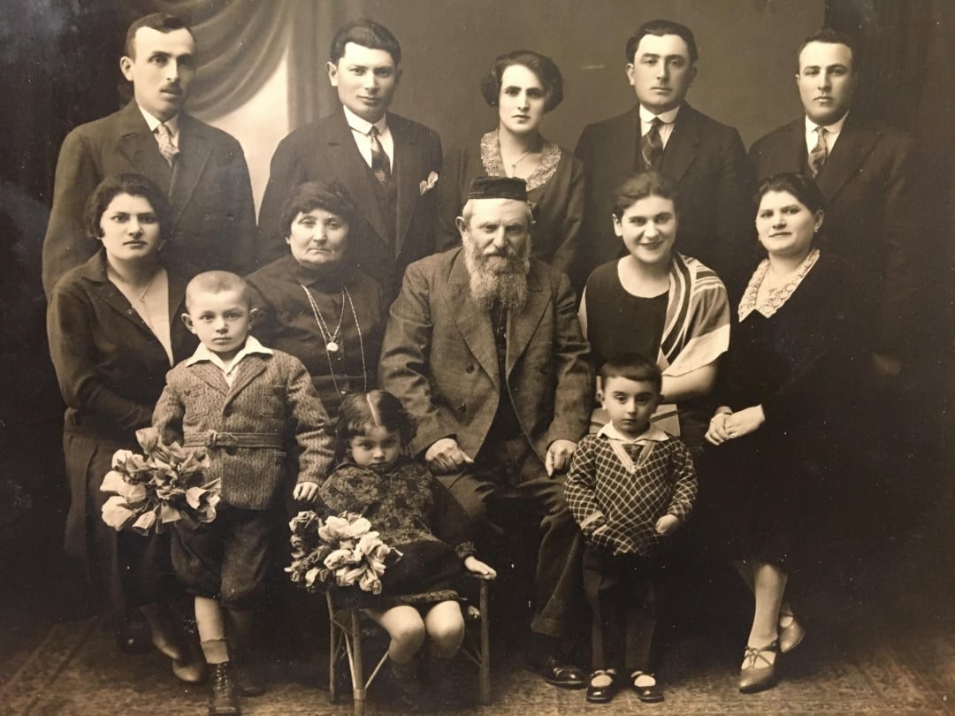 A sepia-toned vintage family portrait featuring eleven adults and three children. The adults are dressed formally, and two of the children hold bouquets while sitting in the front row. Everyone faces the camera and appears serious.