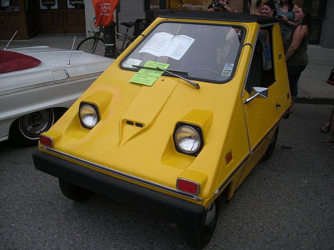 A small, yellow, vintage electric car with a boxy shape is parked on a street during a public event. Papers are visible on its windshield, and a group of people stand nearby.