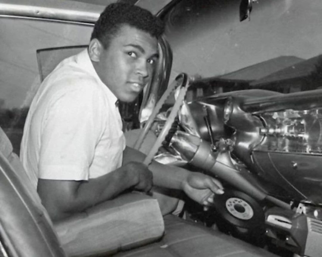 A young man sits in the driver's seat of a vintage car, looking toward the camera while holding the steering wheel. The car's interior and dashboard are clearly visible in the black and white photo.