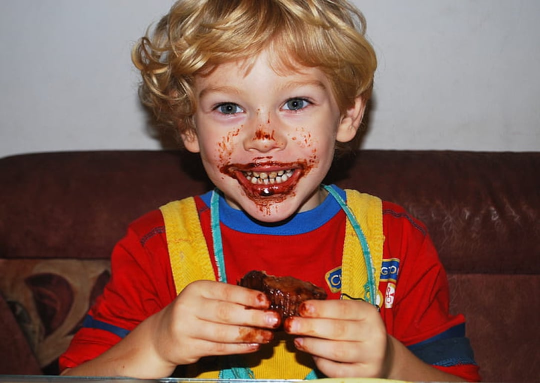 A young child with curly blonde hair and a red shirt smiles widely, their face and hands smeared with chocolate, while holding a piece of chocolate cake.