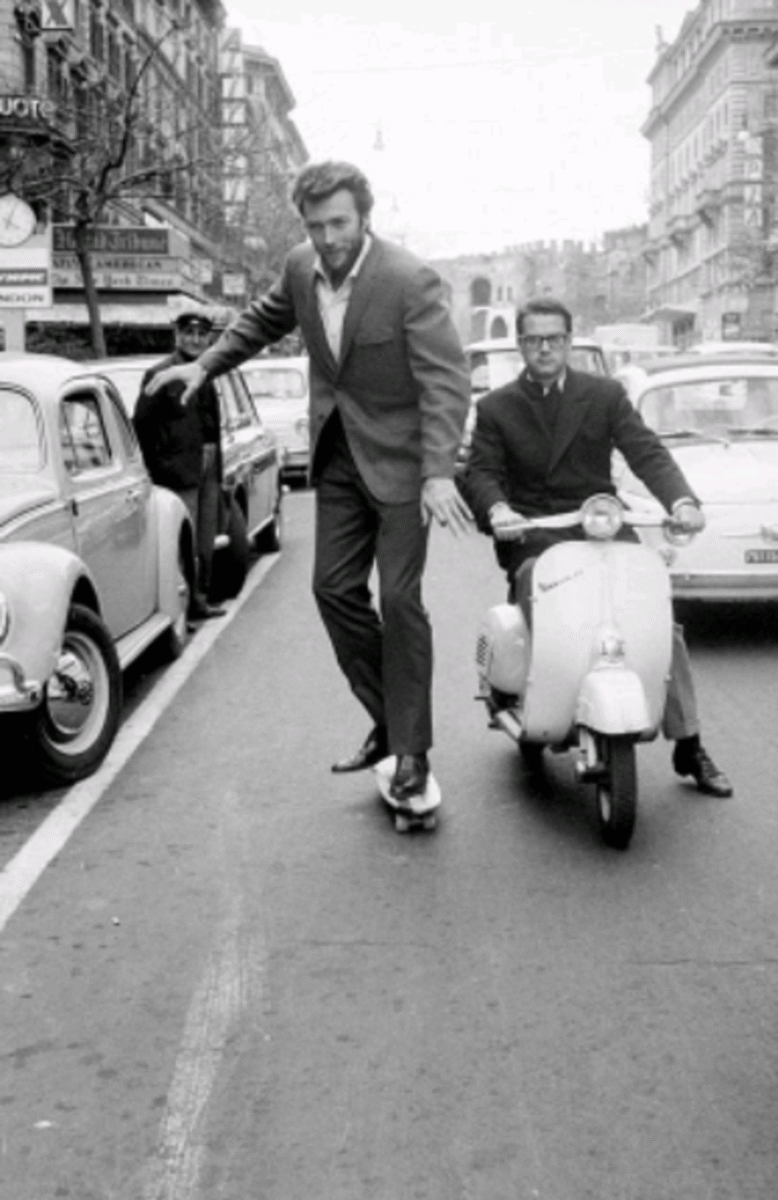 A man in a suit skateboards down a busy city street while another man in glasses rides a scooter behind him. Cars are parked along the sides, and buildings line the street in the background. The image is in black and white.