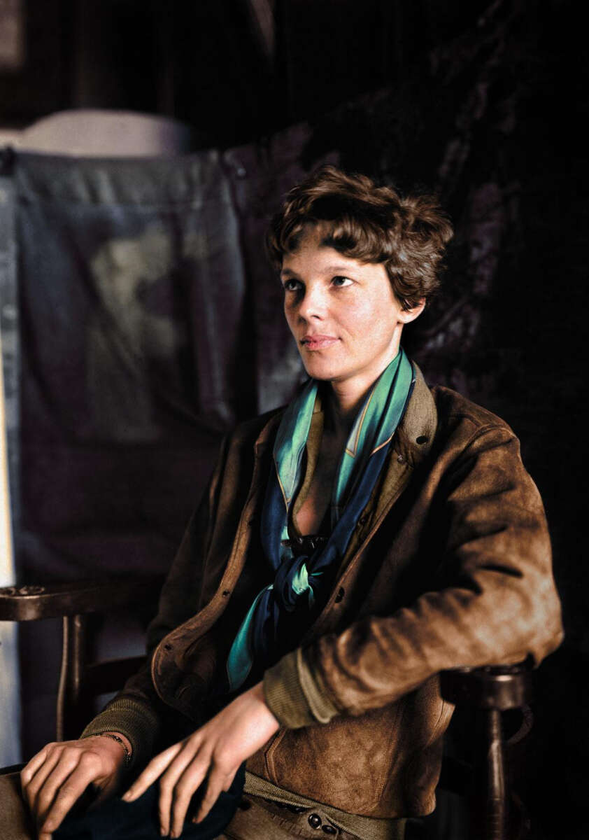 A woman with short, wavy brown hair sits in a chair wearing a brown jacket and a blue-green scarf, looking thoughtfully to the side against a dark, blurred background.