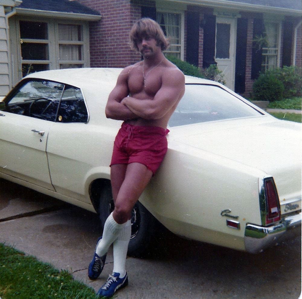 A muscular man with a mustache, wearing red shorts, white knee-high socks, and blue sneakers, stands shirtless with arms crossed, leaning against a vintage cream-colored car in a suburban driveway.
