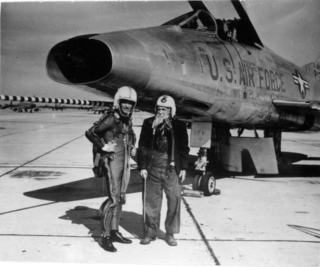 Two people in flight suits and helmets stand beside a U.S. Air Force jet on an airfield. The jet’s nose is prominent, and both individuals appear to be conversing or posing for the photo.