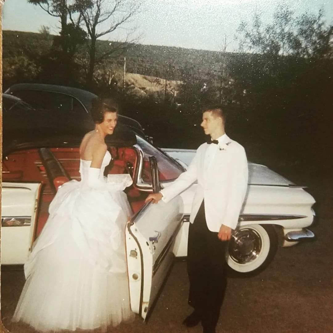 A young woman in a white prom dress stands by a classic white car as a young man in a white tuxedo jacket and black pants holds the car door open for her. They are outdoors, smiling at each other in the evening light.