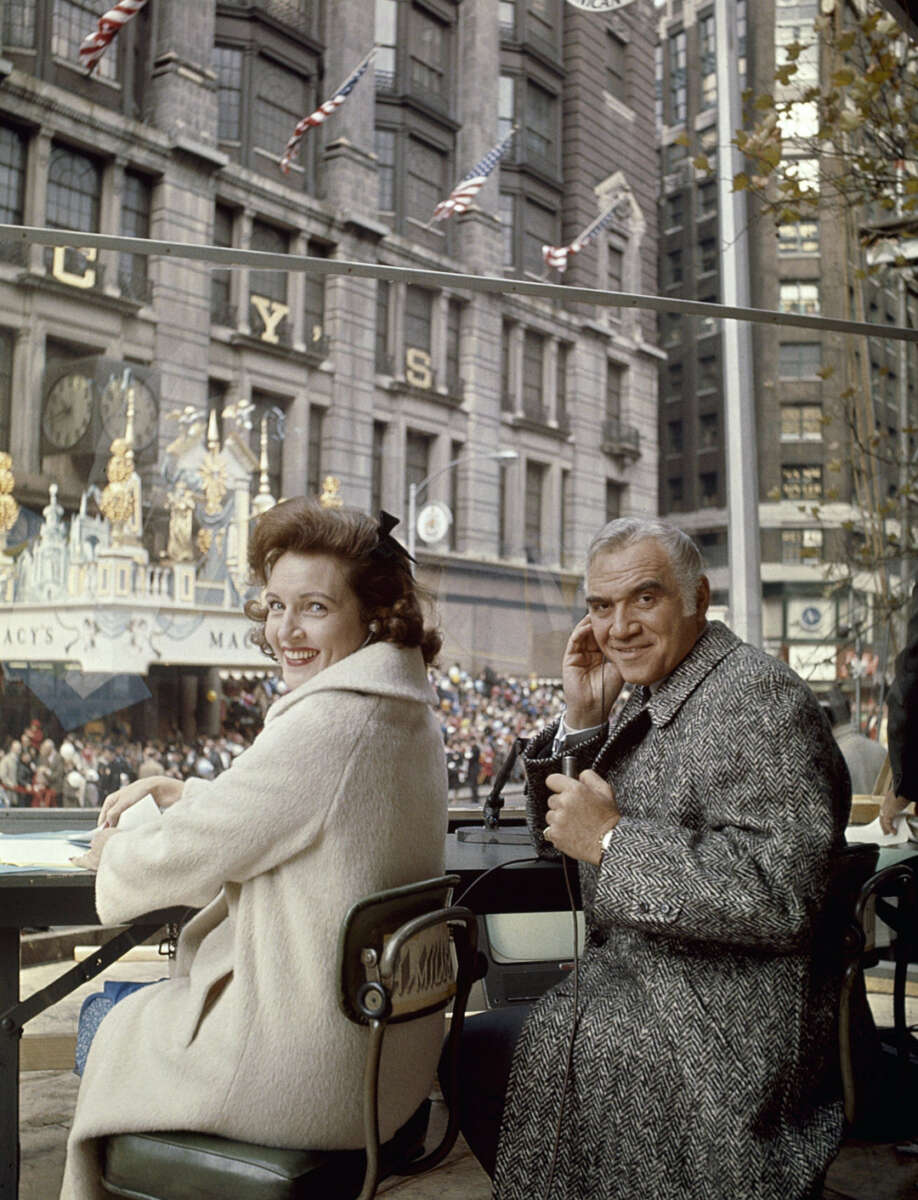 A woman and a man in coats sit at a table with microphones, smiling at the camera during a parade. Macy’s department store and festive decorations are visible in the background.
